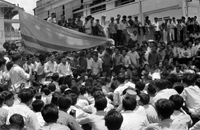 "Unidentified", a large crowd of young Vietnamese men, sit outside watching a man give a speech . A large South Vietnamese flag hangs behind him. In the background, more men stand behind the speaker and sit on a wall . This is likely a part of the anti-Diệm student protests.