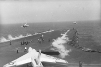 Men working on an aircraft carrier in open water. One Naval plane is still on the runway. Three have just taken off.