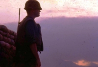 An American soldier standing near a sandbag wall at either dusk or dawn.