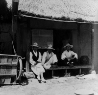 "Korea-Negs 181-3(1)-182-3(1) 13.3 Korean Men" Three old Korean men in traditional clothes sit on the porch of a traditional house. A handcart sits at the far left of the frame. From scrapbook page 23.