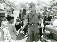 An American soldier named Hibbs is about to shake a young child's hand as other villagers watch.
