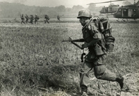 "Phuoc Tuy Province, Vietnam - Paratroopers of the 173rd Airborne Brigade dash for helicopters of the 82nd Aviation Battalion during the air assault phase of recent search and destroy operations in Phuoc Tuy Province." An American soldier runs through a field. Another group of soldiers and two helicopters are in the background.