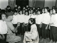 "Saigon - TET Envelopes - Chaplain (Colonel) Theodore V. Koepke, Chief of Chaplains, U.S. Military Assistance Command, Vietnam, holds the TET Envelopes, containing donations by the NACV Chaplains Corps, for the girls in the Buddet Institute Orphanage in Saigon. Assisting Chaplain Koepke in distributing envelopes is Miss Lee Eckles, of San Francisco, California, came to Vietnam at her own expense, with the assistance of friends, to try to help the people of this country in their struggle for freedom and independence." An American man in military uniform and a woman wearing an ao dai crouch amongst a group of Vietnamese girls.  The man holds something in his hands, possibly envelopes.