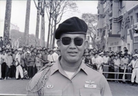 A Vietnamese soldier named Nhon looks at the camera. There is a crowd behind a barricade in the background.