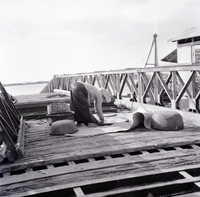 A Vietnamese woman appears to be threshing rice on a wooden dock or bridge. She is surrounded by baskets.