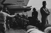 "7th Fleet" An American man wearing a headset and two other men near what may be artillery on the USS Kitty Hawk. In the foreground stands an officer giving the thumbs-up sign.