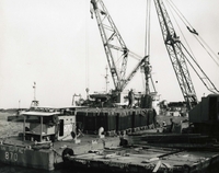 "Cargo Delivery - Pier Team #16 offloads LCM-8 #870 at Museum Annex pier within the city of Da Nang. The cargo consists of beverages bound for the Army-Air Force Exchange. Just 95 minutes earlier it was in the hold of a merchant vessel anchored in Da Nang harbor." A Naval dock with large cranes and crates of cargo.