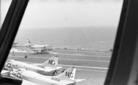 Four Naval planes on an aircraft carrier in open water. Photo taken from the window of another plane.