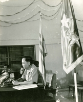 Nguyá»…n KhÃ¡nh appears to be at a press conference. He sits at a desk with microphones in front of him. (damaged)