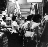 "Sarawak." A crowd of fashionable women stand in a narrow market aisle. On both sides the stalls appear to be selling fabrics. A man selling the fabric is on the far left of the photo. From scrapbook page 13.
