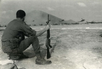 "Suoi Da, Vietnam -A 173rd Airborne Brigade trooper watches the aerial acrobatics of the U.S. Army resupply choppers Heli lifting combat cargo to Operation 'Junction City' soldiers sweeping through War Zone 'C.' The month-long sweep and clear operation is the largest of the conflict in Vietnam, with the Army's multi-division force adding more than 1,100 'kills' to the 'Junction City' sweep." An American soldier crouches holding a gun. In the background, a large group of helicopters take off.