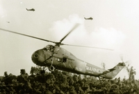 "The UH34D is a single engine helicopter designed for transportation of cargo and personnel including aeromedical evacuations." A US Marine Corps helicopter in flight above a jungle. There are two more helicopters far in the background.