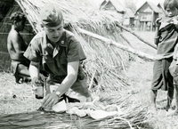 An American soldier named Hibbs examining a newborn baby. He holds a bottle of medicine. Villagers watch in the background.