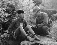 A Vietnamese prisoner sitting next to an American soldier, as the soldier is reading something to him. The prisoner has a piece of paper tied around his neck and his feet are shackled.