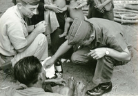 Two American soldiers examine and provide medical care to an injured villager's foot. One of them is smoking a cigarette.