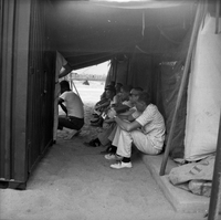 A group of American men are sitting underneather a tent watching something off camera.