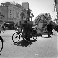 "Macau" Men are walking down the street in a parade or processional with rickshaws decorated with shrines and flowers.
