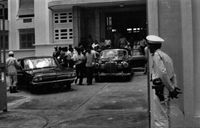 "Unidentified", a group of Vietnamese men are either arriving or leaving from a gated building. Two cars sit in the gated driveway, and several guards stand nearby.