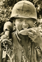 American soldier is seen taking a bite out of a corn cob. He is holding something else (possibly a missile) in the other hand, and appears to be posing for the picture.