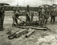"In Our Hands - Parts from four 75mm "Pack" howitzers, the first artillery pieces captured by U.S. forces in Vietnam, are displayed at a 9th Infantry Division fire support base. The weapons, discovered 30 miles east of Saigon on Oct. 14, were found near a tunnel complex that contained more than 500 enemy weapons." A group of American soldiers stand around a pile of broken machines and weapons.  In the background there is barbed wire, a sandbag wall, and tents.