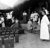 "Korea-Negs 183-1(2)-184-1(2) 10.Farm" A group of Korean people stand in a courtyard of a traditional farm, looking at the camera. The courtyard appears to be filled with drying bricks. Most of the people pictured are women and children in traditional clothes, though there is one man in modern western dress. From scrapbook