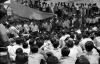 "Unidentified", a large crowd of young Vietnamese men, sit outside watching a man give a speech . A large South Vietnamese flag hangs behind him. In the background, more men stand behind the speaker and sit on a wall . This is likely a part of the anti-Diệm student protests.