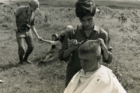 "Pleiku, Vietnam (Army IO) - Army PFC Stanley Crump, Winthrop, N.Y., gets his hair cut to meet Airborne standards, while in the background, another trooper pays the pretty cashier for his haircut." A Vietnamese man cuts an American soldier's hair. In the background, an American soldier hands a Vietnamese woman something while she sits on the ground.