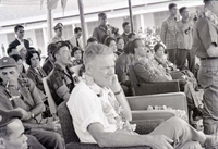 A seated Henry Cabot Lodge Jr is wearing a lei. He is surrounded by many other military members and officers, from both the Vietnamese and American militaries.