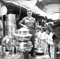 "Ca Mau" American soldier standing behind a stall of cooking pots with children standing beside him for the photo.