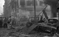 "Embassy Bombing" Two cars have collided in the steaming rubble. A crowd of people stands nearby to take pictures. Two bodies lay in the rubble beside the crowd.