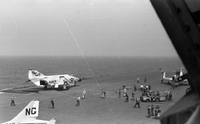 Men working on an aircraft carrier in open water. Three Naval planes are in sight. Photo may have been taken from within a fourth plane.