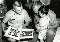 Two American soldiers are looking through a photo album with a young Vietnamese child. One soldier is holding the album as the other kneels behind the child.