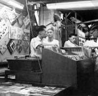 "Sarawak." A stall in a Sarawak night market selling LP records and some printed materials (possibly small books). There are 3 vendors and 1 customer in the photo. The stall is decorated with LP covers and there is a record player behind the case. From scrapbook page 13.