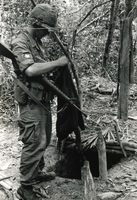 "Bien Hoa, Vietnam - Specialist 4 Homer A. Duncan of Atlanta, Ga., takes wash off the line in a Viet Cong camp after the VC had fled from the U.S. Army Airborne troops during recent operations by the 173rd Airborne Brigade in War Zone 'D'." An American soldier stands over a tunnel entrance in a forest. He holds a gun and a piece of cloth.