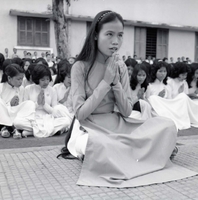 A Vietnamese woman is sitting on the ground and praying. Many other women are seen praying behind her in rows.