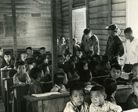 "New School at Le Phung - U.S. Army Special Forces advisor SFC Charles R. Smith (of Okmulgee, Okla.) visits school at Le Phung, a village of 800 about 250 miles NNE of Saigon, close to the Cambodian border. A few months ago Le Phung was a Viet Cong village, before Special Forces advisors and CIDG troops from Duc Co surrounded it and drove out VC. Sergeant Smith was one of the advisors who liberated Le Phung. Now he checks on its progress, visiting the new school. At his left is Dihn Tri Vien, interpreter for "A" Team 215. Schoolmaster (in white shirt) is 16 years old." An American airman and a Vietnamese soldier are in a classroom full of children.  They appear to be looking at one child's work.  The classroom is bare, with rows of school desks and benches.