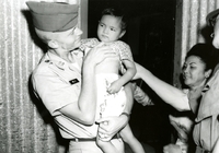 An American soldier holding a young Vietnamese child, with another soldier and Vietnamese woman in the background.