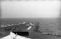 Men working on an aircraft carrier in open water. Two Naval planes are still on the runway. Two have just taken off.