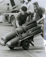 "Ready the Bombs - Aviation Ordnance men aboard the Seventh Fleet attack aircraft carrier USS Kitty Hawk roll 500-pound bombs across the flight deck to attack and fighter aircraft just prior to their launch against targets in North Vietnam." Two American men use a cart to move three missiles, probably onboard an aircraft carrier.