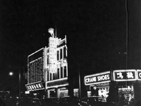"Sarawak." View of a Sarawak street at night. There are cars on the road and buildings lit with flourescent signs and bright shop windows. One business is labelled "Crane Shoes." The other signs are in Chinese characters. One building appears to be a jewellry store, since it has a ring on its sign. From scrapbook page 13.