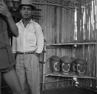 "Montagnards - Showplace Strat. Ham - 8-21-62 - In MTs - Faces - Blockhouses" A nicely dressed Vietnamese man is standing near some medical supplies in a hut.