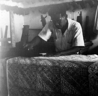"Indonesian Arts and Crafts Negs [Negatives]." An Indonesian man stamps wax onto fabric to make batik. There is wooden furniture stacked against a wall in the background. From a scrapbook page 10.
