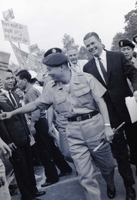 Robert McNamara walks behind Nguyá»…n KhÃ¡nh in a crowd. Henry Cabot Lodge, Jr., walks behind them.