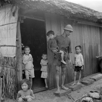 Original title: "VN Life", A man stands with five small children in the doorway of a traditional wood home with a tatched roof. There appears to be a pile of coconut husks in front of the house.
