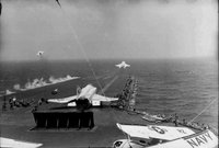 American men working on an aircraft carrier in open water. Two Naval planes are still on the runway, one with detached wings. Two others have just taken off.