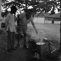 Original title "Cao Lanh - XON + MAAG ", 2 men cooking in a yard, a tree and some houses in the background