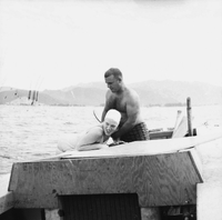 An American woman is leaning over on a boat as an American man stands behind her.  They are dressed in swim suits.