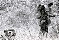 Two American soldiers are in a field. One is laying down as the other stands nearby.