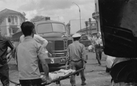 "Embassy Bombing" Two men carry a body on a stretcher toward an ambulance. Other people and cars crowd the street.