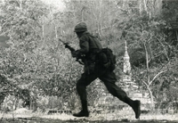 "Phuoc Tuy Province, Vietnam - An Army paratrooper of Company A. 1st Battalion, 503rd Infantry moves out under fire to flank Viet Cong located on high ground to his right. Assault up the hill by this unit forced the VC to withdraw." An American soldier runs through the forest. There are two stupas in the background.